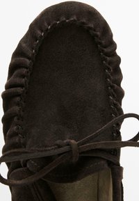 Mocassino in suede marrone scuro con cuciture visibili e un laccio in pelle annodato su uno sfondo chiaro.