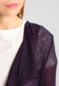 Cardigan di maglia leggero viola che scivola su una maglietta bianca, con colletto morbido e tessuto strutturato con sezioni trasparenti.