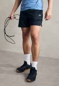 Svarte trening shorts med sidelukking, sammen med svarte joggesko og hvite ankelsokker. Individet holder et hoppetau, iført en blå topp.