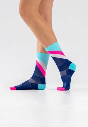 Legs wearing ankle socks with bold navy, turquoise, white, and pink geometric patterns, standing on a white surface with one foot slightly raised.