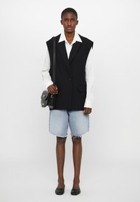Black vest with shoulder pads and single button, white shirt beneath, paired with blue denim shorts and black flats. Minimalistic design.
