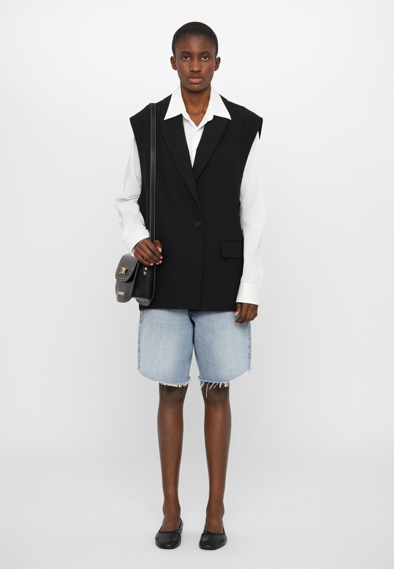 Gilet nero con spalline e un solo bottone, camicia bianca sotto, abbinato a pantaloncini in denim blu e ballerine nere. Design minimalista.