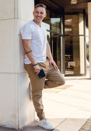 White short-sleeve v-neck shirt, beige cargo pants, white sneakers, holding a dark phone. Leaning against a light-colored wall.