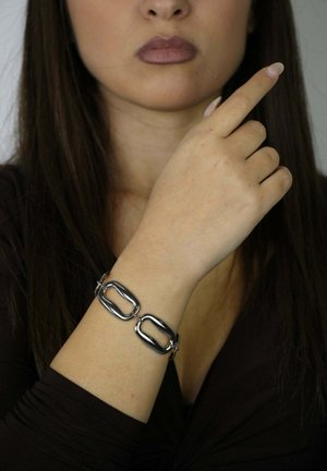 Eine Frau, die ein dunkles Oberteil trägt, zeigt ein silbernes Gliederkettenarmband an ihrem linken Handgelenk, während ihre rechte Hand nach oben zeigt.