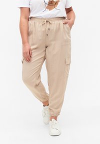 Beige cargo kalhoty s elastickými manžetami a bočními kapsami, vyrobené z lehkého materiálu. Model je kombinuje s bílým tričkem a teniskami.