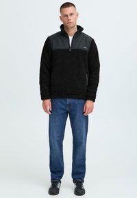 Svart fleecepulle med kvart glidelås, har en glatt svart panel på brystet og grå detaljer, kombinert med blå denimjeans.