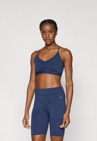 Μπλε ναυτικό αθλητικό τοπ crop με ρυθμιζόμενους ιμάντες και ραμμένες λεπτομέρειες, συνδυασμένο με σορτς ποδηλασίας σε ψηλόμεση γραμμή στο ίδιο χρώμα. Ύφασμα λείο και ελαστικό.