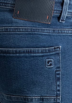 Nahaufnahme der Gesäßtasche einer blauen Jeans mit gesticktem Markenlogo und einem schwarzen Lederetikett über der Tasche.