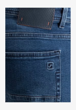 Zbliżenie na tylną kieszeń niebieskich dżinsów z przeszytym logo marki i czarną skórzaną łatą nad kieszenią.