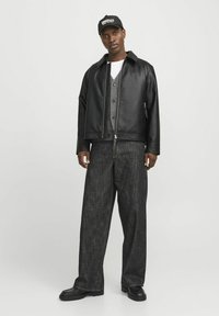 Black leather jacket with a zip, worn over a gray cardigan and white t-shirt. Loose-fitting dark denim pants and black shoes complete the look.