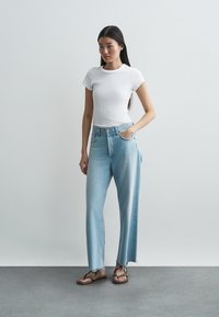 Blugi largi, de culoare albastru deschis, cu tivuri franjurate, asortați cu un tricou alb mulat. Modelul stă pe o suprafață gri, pe fundal simplu.