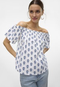 Hvid off-shoulder top med blå blomster mønstre, korte ærmer og rynket halsudskæring. Blødt stof; afslappet pasform. Modelleret med denim bukser.