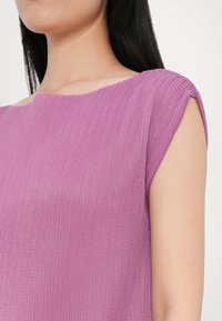 Lilla ribbet topp med vid halsutskärning och korta ärmar, med en strukturerad tyg och enkel design, placerad mot en ljus bakgrund.