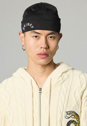 Giovane che indossa un berretto nero con scritta, orecchini a cerchio argentati e una felpa con zip crema a maglia a trecce con una toppa a forma di serpente sul petto.