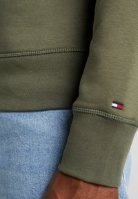 Sudadera verde oliva con puños de canalé, que presenta una pequeña etiqueta de logotipo roja, blanca y azul en la manga, combinada con denim azul claro.