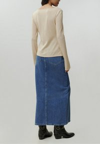 Beige Langarmshirt, kombiniert mit einem blauen Jeans-Maxirock mit zwei Gesäßtaschen. Das Model trägt schwarze Knöchelstiefel.
