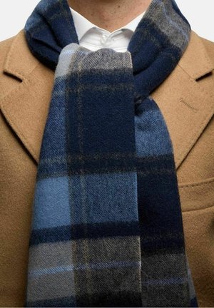 Ullscarf med ett blått och marinblått rutmönster, mjuk textur och fransade kanter, hängande runt halsen mot en beige kappa.