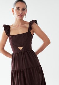 Bordeauxrode jurk met een vierkante halslijn, gerimpelde bandjes en een uitgesneden voorkant. De stof is gestructureerd en heeft een gelaagde, uitlopende rok.