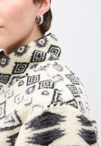 Flis jakna s crnim i sivim geometrijskim uzorcima na krem pozadini; srebrna minđuša u obliku obruča na lijevom uhu.