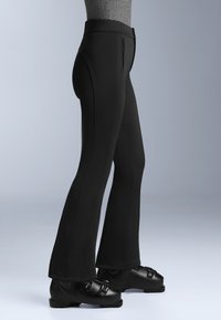 Černé lyžařské kalhoty vyrobené z texturované látky, s vysokým pasem, švy po přední části a přiléhavými rozšiřujícími se nohavicemi, doplněné černými lyžařskými botami.