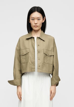 Frau mit glatten schwarzen Haaren trägt eine kurze khakifarbene Jacke mit großen Fronttaschen über einem weißen Kleid.