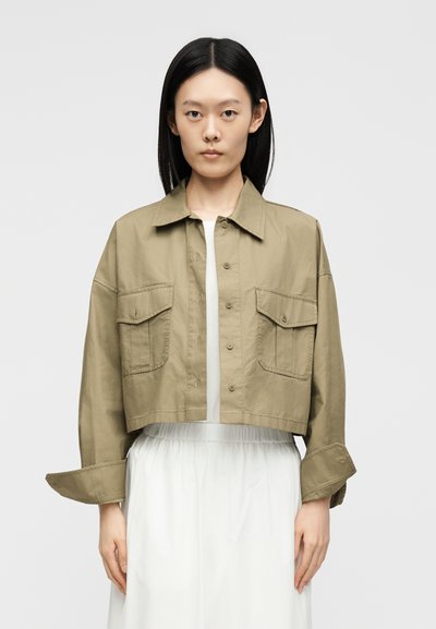 Woman with straight black hair wears a cropped khaki button-up jacket with large front pockets over a white dress.