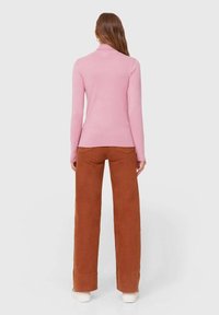 Nainen seisoo selin, päällään vaaleanpunainen pitkähihainen paita, rustinväriset leveälahkeiset housut ja valkoiset kengät yksinkertaisella taustalla.