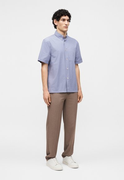 Άνδρας που στέκεται, φοράει ανοιχτό μπλε πουκάμισο με κοντά μανίκια και κουμπιά, καφέ παντελόνι με ίσια μπατζάκια και άσπρα αθλητικά παπούτσια σε απλό φόντο.