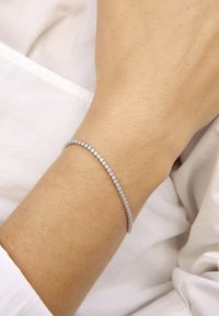 Muñeca con una delicada pulsera de tenis de plata con pequeños diamantes redondos, sobre un fondo de manga de camisa blanca.