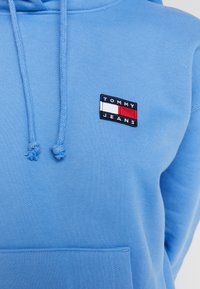 Ljusblå hoodie i mjukt material med dragsnören. Har ett litet broderat logopatch med texten "TOMMY JEANS" i rött, vit och blått.