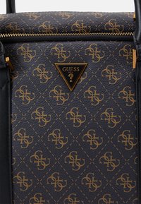 Schwarze synthetische Handtasche mit geprägtem goldenen Logo-Muster, ausgestattet mit einem dreieckigen goldenen Anhänger mit "GUESS" und einem Reißverschlussverschluss oben.