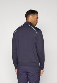Veste zippée marine avec col haut, ornée de surpiqûres blanches aux épaules. Tissu lisse et design ajusté.