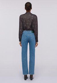 Frau mit geflochtenem Dutt, die eine transparente schwarze Bluse mit Tiermuster, blaue hochgeschnittene Jeans und schwarze Absatz-Ankle-Boots trägt, mit dem Rücken zur Kamera.