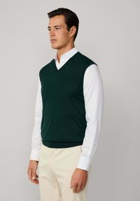 Ciemnozielony sweter w serek z fakturowanej dzianiny, noszony na białej koszuli z kołnierzykiem, w połączeniu z jasnymi spodniami.