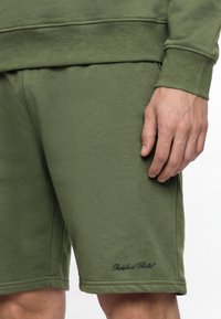 Homme portant un short vert olive et un sweat assorti, montrant une main reposant sur le côté avec un petit texte noir brodé sur le short.