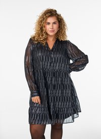 Musta mekko, jossa on läpikuultavat pitkät hihat, V-aukko, nappidetaili ja kerroksittain leikattu hame, jossa on hopeisia viivakuvioita koko mekossa.