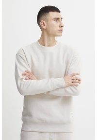 Világos bézs színű pulóver, puha textúrájú anyagból, kerek nyakkivágással, bordázott mandzsettákkal és logós címkével az alján. Egyszerű, minimalista design.