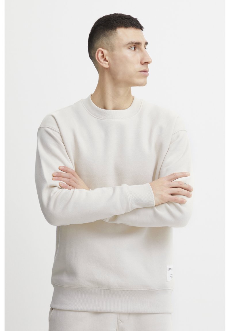 Világos bézs színű pulóver, puha textúrájú anyagból, kerek nyakkivágással, bordázott mandzsettákkal és logós címkével az alján. Egyszerű, minimalista design.