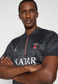 Fekete sportmez texturált, függőleges csíkokkal, piros részletekkel és logókkal, többek között Jordan emblémával és a Paris Saint-Germain címerrel.