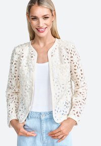Cardigan in pizzo color avorio, con motivi floreali intagliati, scollatura rotonda e chiusura con bottoni. Sotto, è visibile una canotta bianca.