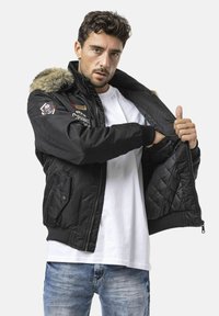 Schwarze Bomberjacke mit einem mit Fell gefütterten Kapuzen, Reißverschluss und seitlichen Taschen. Verziert mit Stickereien und gesteppter Innenfütterung. Wird über einem weißen T-Shirt getragen.