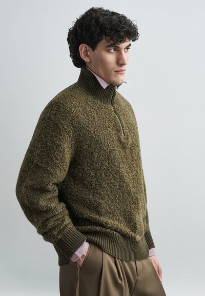 Olijfgroene trui met een hoge kraag en halve ritssluiting, gemaakt van gestructureerde stof. Draagt boven een roze overhemd met kraag, gecombineerd met beige broek.