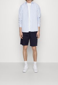 Chemise à rayures bleu clair et blanche avec boutons, portée sur un t-shirt blanc, associée à un short marine et des baskets blanches. Design simple, coupe décontractée.
