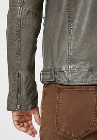 Veste en cuir gris avec une finition texturée, dotée d'un poignet zippé et d'un détail à bouton-pression à la taille, associée à un pantalon en tissu marron.