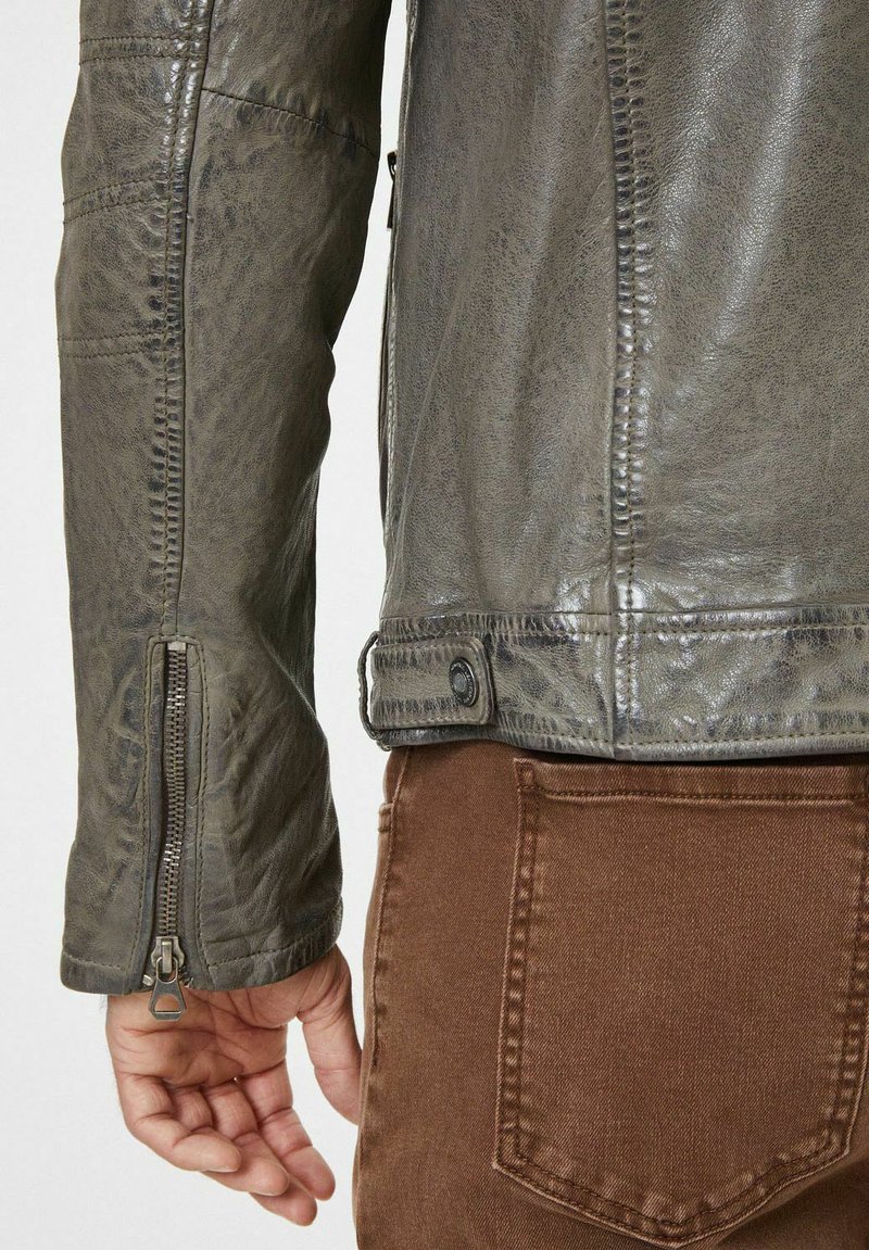Veste en cuir gris avec une finition texturée, dotée d'un poignet zippé et d'un détail à bouton-pression à la taille, associée à un pantalon en tissu marron.