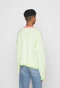 Zielono-biały sweter dzianinowy w falisty wzór z różowym ściągaczem przy dekolcie i mankietach, charakteryzujący się luźnym fasonem i opadającymi ramionami.