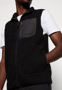 Muž v černé fleecové vestě se zapínáním na zip a náprsní kapsou označenou "Originals" přes bílou košili s krátkým rukávem, s rukou v kapsě.