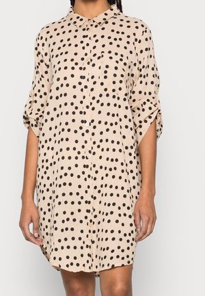Personne portant une robe-chemise beige ample à pois noirs et manches retroussées, debout devant un fond uni.