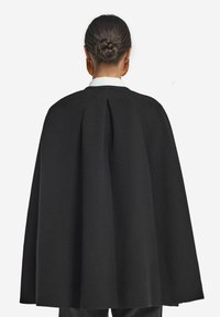 Femme aux cheveux bruns dans un chignon torsadé, portant de grandes boucles d'oreilles et un vêtement de style cape noir par-dessus une chemise à col blanc, vue de dos.