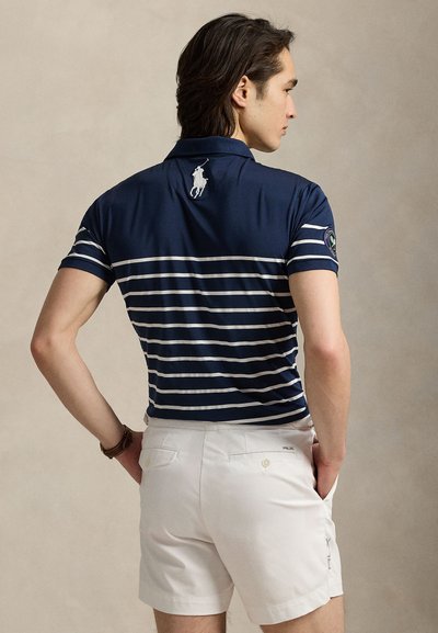 Čovjek okrenut leđima, odjeven u tamnoplavu polo majicu s prugama i bijele kratke hlače, ruke u džepovima, stoji ispred neutralne pozadine.